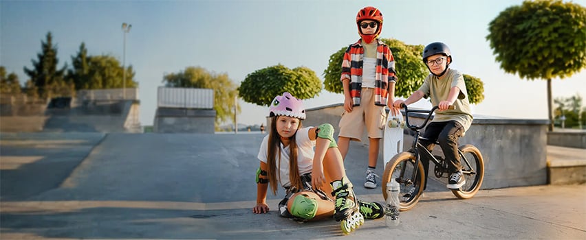 Troje dzieci w kaskach i sprzęcie ochronnym Wozinsky na deskorolce, rolkach i rowerze BMX na skateparku
