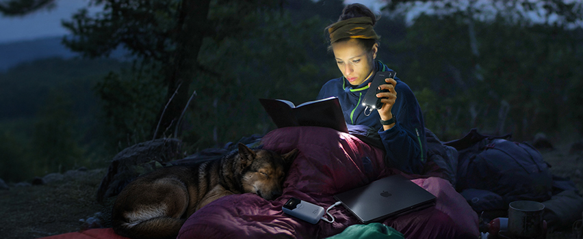 Kobieta czyta książkę w namiocie z psem i ładuje smartfon i laptopa powerbankiem Wozinsky oraz latarką czołową